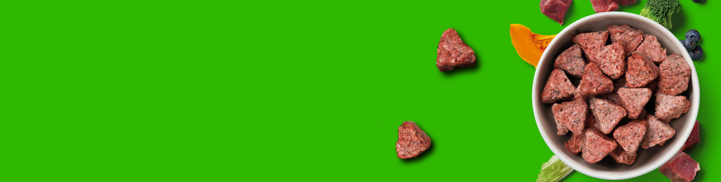 A bowl of heart-shaped pet food sits on a bright green background, surrounded by vegetables, blueberries, and pieces of meat. A few pet food pieces are scattered outside the bowl.