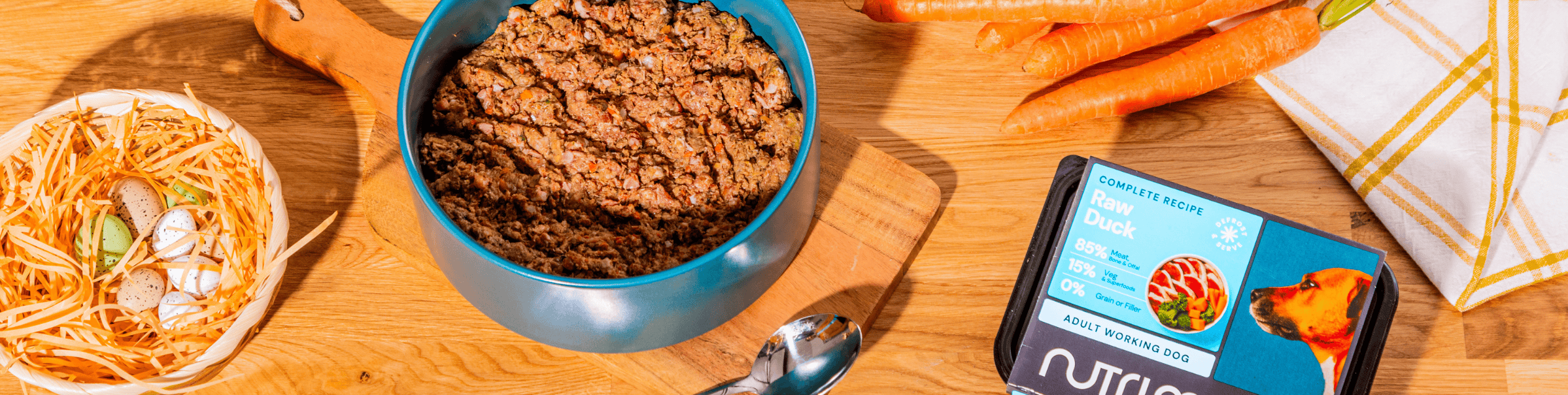 Person serving raw dog food to a brown dog on a kitchen counter. Various packages of Nutriment dog food and chopped carrots are on the wooden surface.