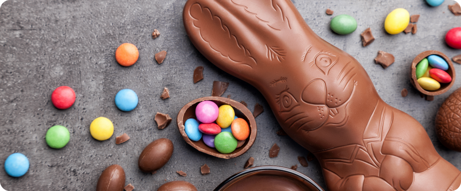 A chocolate bunny with colorful candy pieces and chocolate eggs on a gray surface.