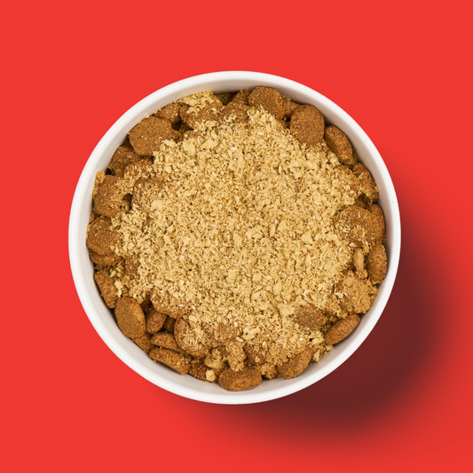 A white bowl of brown kibble with Freeze Dried Topper - Beef 50g sprinkled on top, set against a red background.