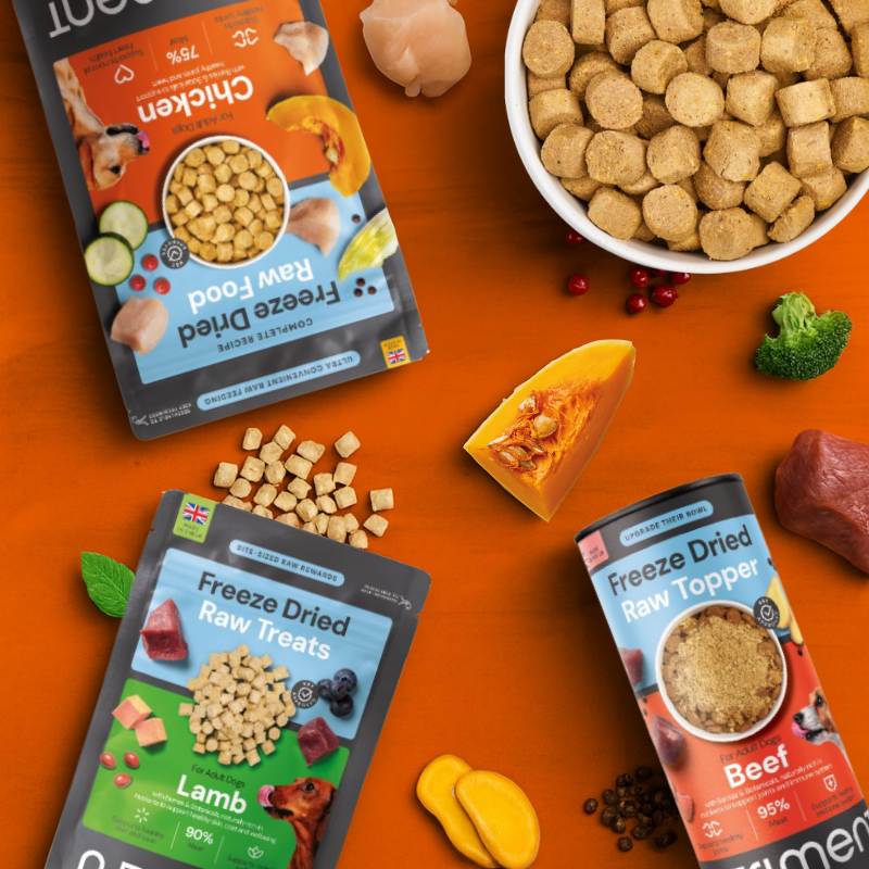 Three bags of freeze-dried pet food and toppers with ingredients and treats scattered on an orange surface.
