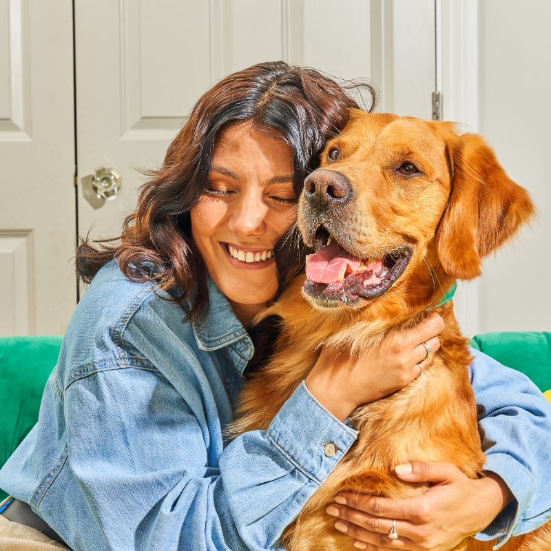 Smiling woman in a denim shirt hugging a happy golden retriever indoors.