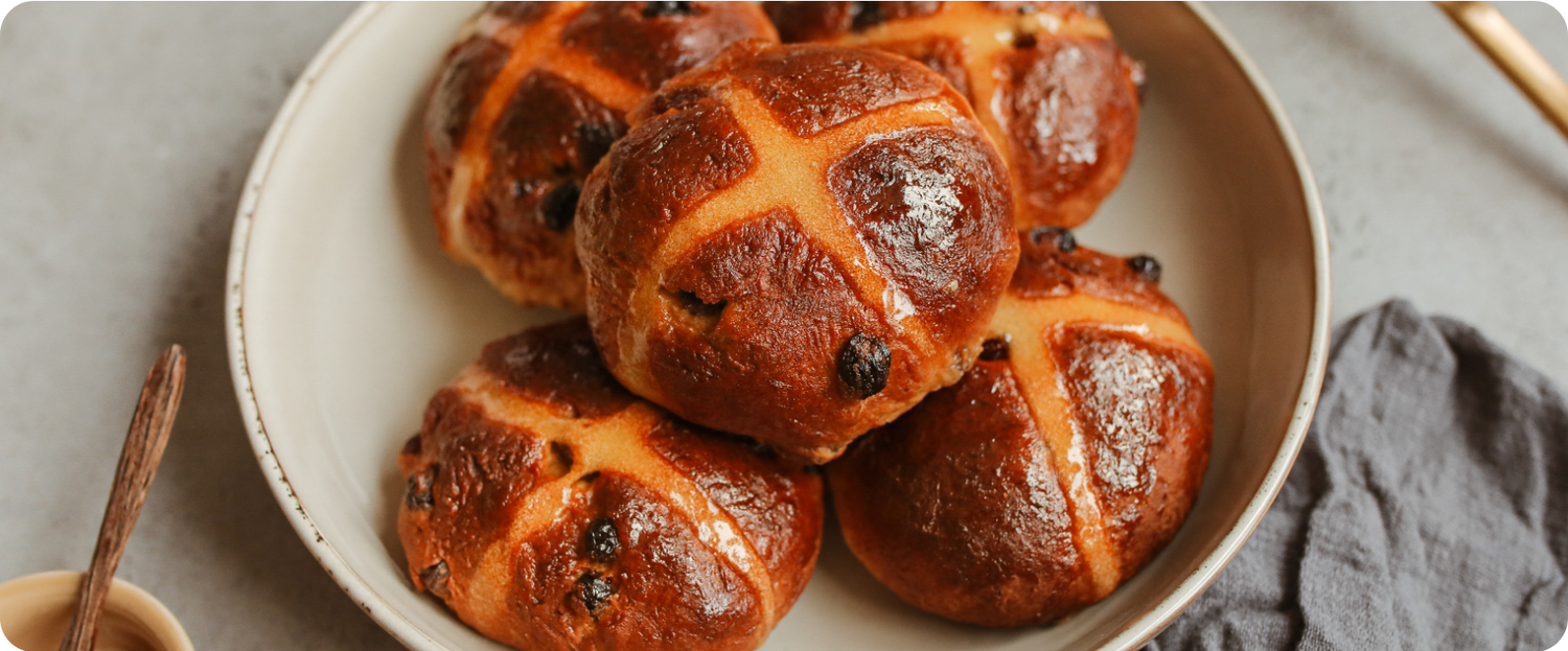 A plate of golden-brown hot cross buns with raisins, marked with a white cross on top.