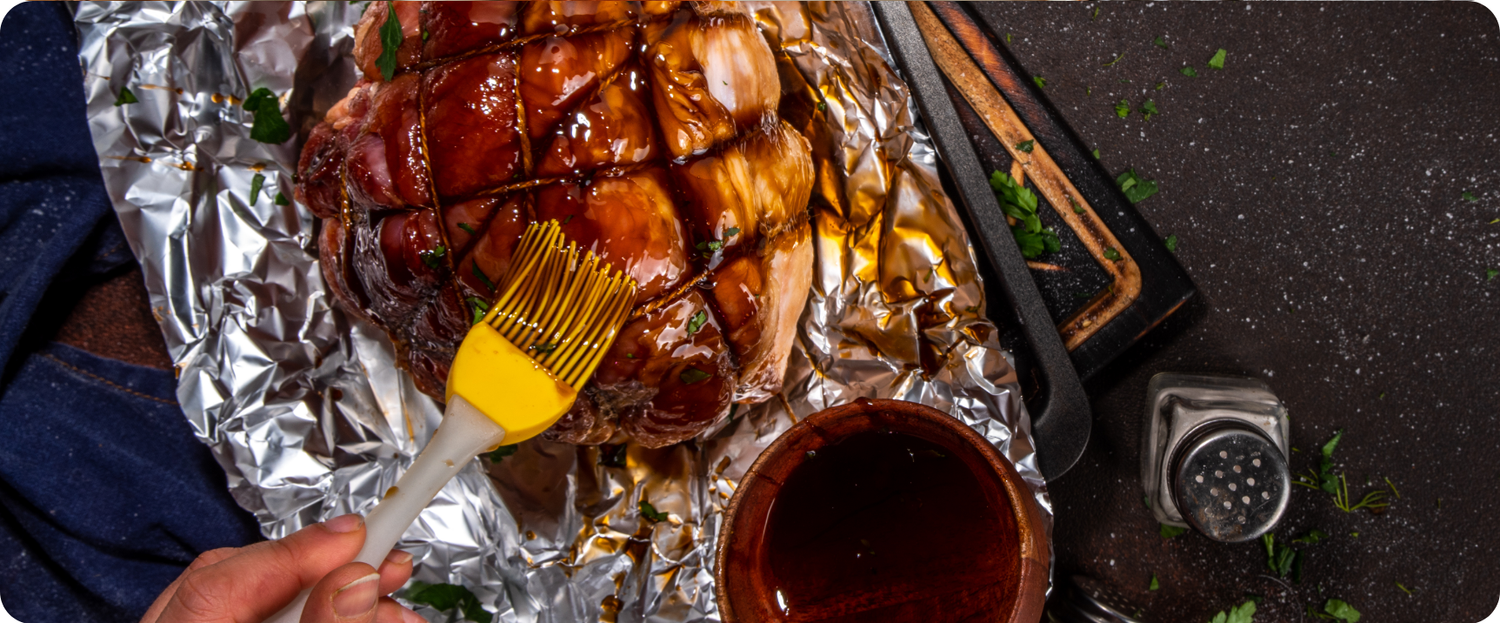 A hand brushes glaze onto a ham resting on aluminum foil, with a bowl of glaze and a pepper shaker nearby.