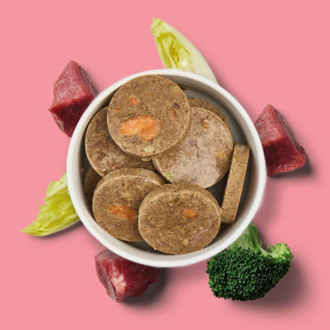 A bowl of Fresh Cooked Medallions - Beef 800g with raw meat, lettuce, and broccoli on a pink background.