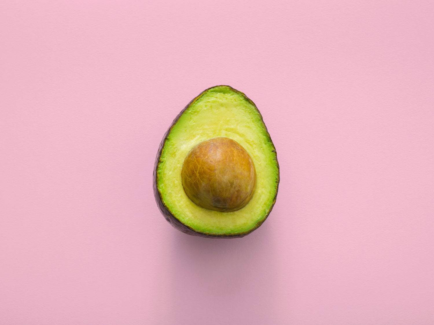 Half an avocado with the pit on a pink background.